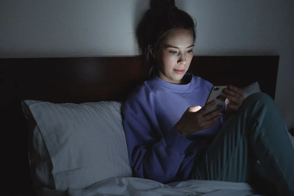 A woman sitting in bed at night looking at a smartphone lit by its screen